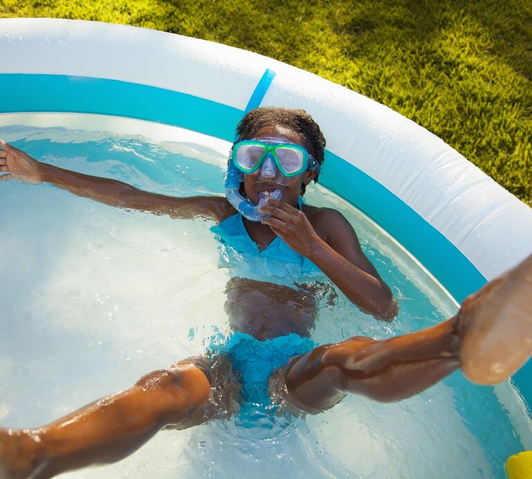Ein fröhliches Mädchen in blauer Badebekleidung und Taucherbrille planscht mit ausgestreckten Armen im aufblasbaren Pool im Garten. | © Getty Images/Kevin Kozicki