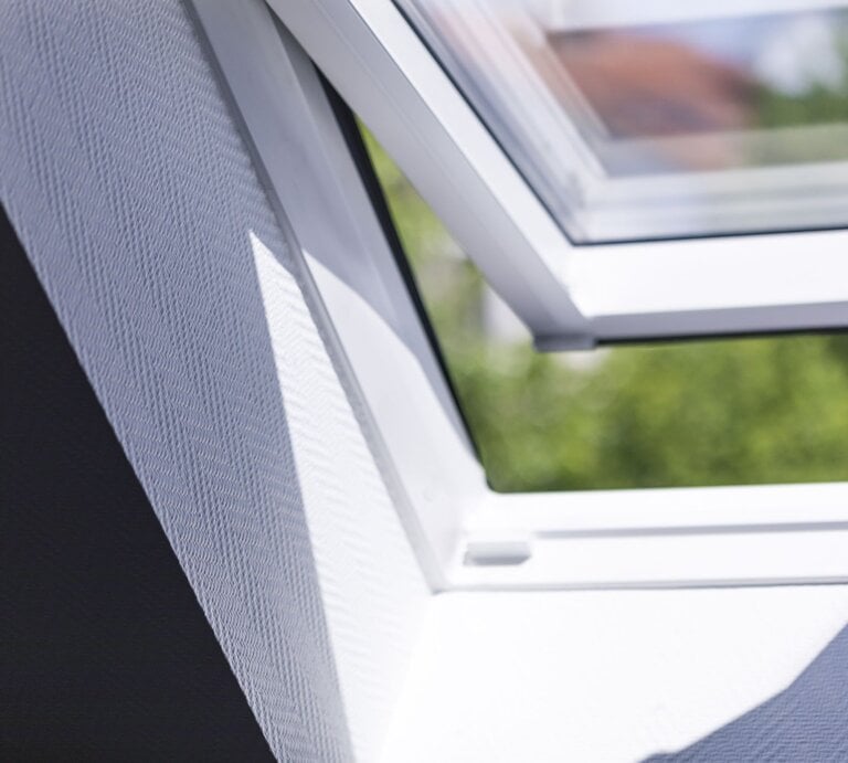 Sonnenlicht fällt durch ein schräges, geöffnetes Dachfenster auf eine weiße, strukturierte Innenwand, im Hintergrund Bäume. | © Getty Images/Malorny