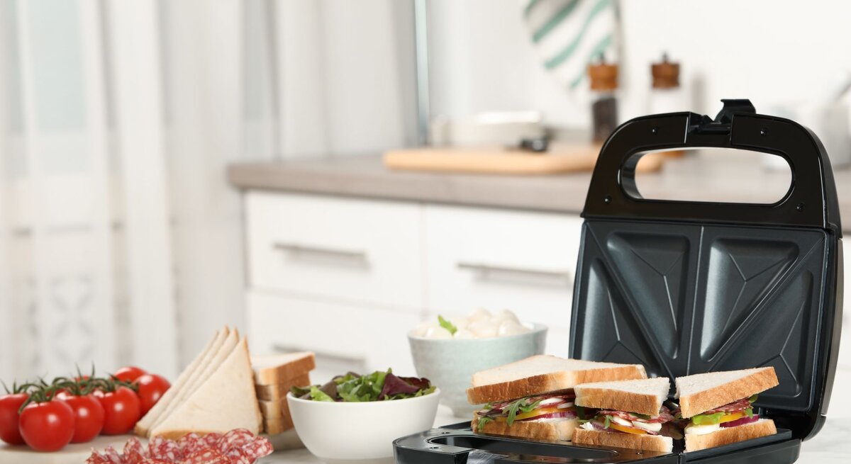 Ein Sandwichmaker mit vier belegten Toasts steht auf einer Küchenarbeitsplatte neben Tomaten, Salami und Schalen mit Zutaten.