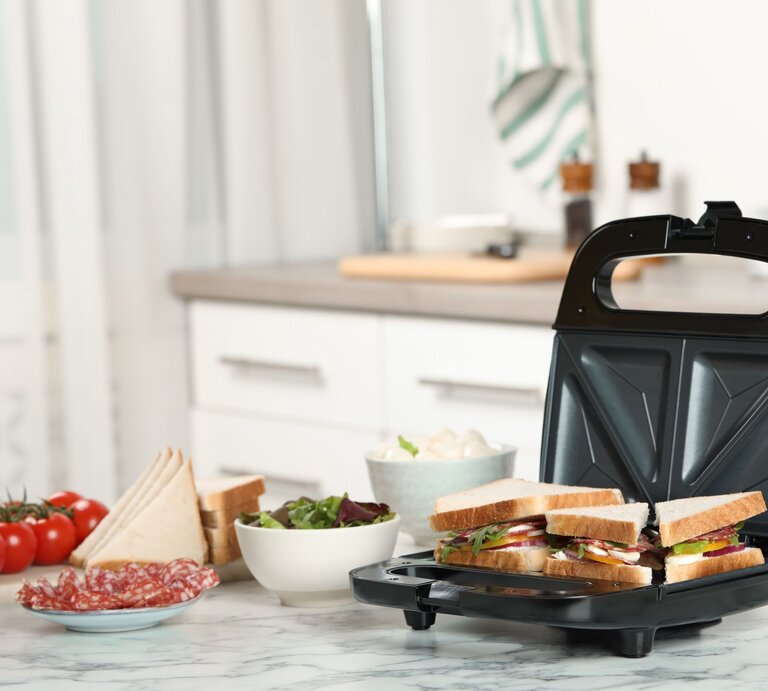 Ein Sandwichmaker mit vier belegten Toasts steht auf einer Küchenarbeitsplatte neben Tomaten, Salami und Schalen mit Zutaten. | © Adobe Stock/New Africa