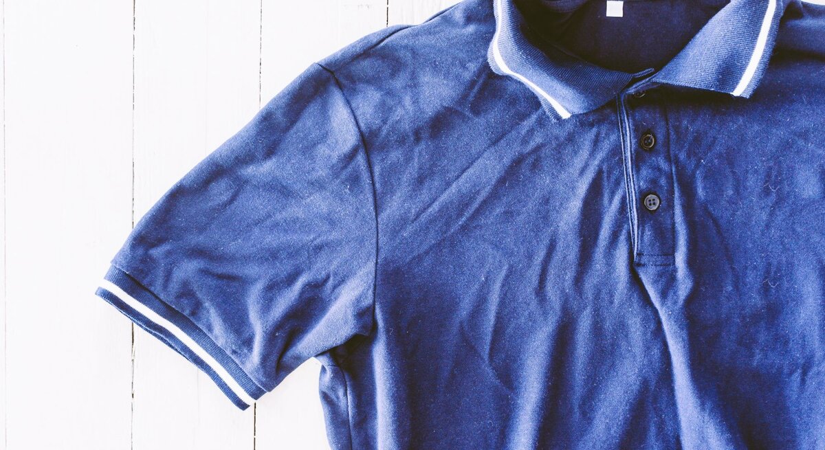 Ein blaues Polohemd mit weißer Zierleiste und drei Knöpfen liegt auf einem weißen Holzboden.