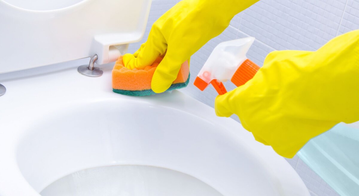 Hände in gelben Gummihandschuhen reinigen mit Schwamm und Sprayflasche eine weiße Toilettenschüssel vor gefliestem Hintergrund.