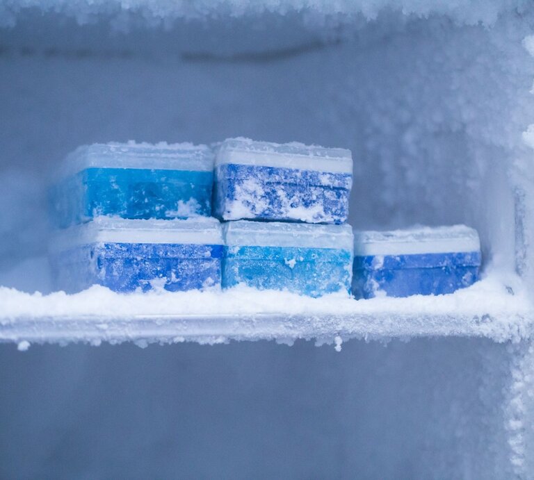 Im frostigen Inneren eines Gefrierschranks liegen übereinander gestapelte, vereiste blaue Kühlakkus. | © iStock.com/jxfzsy