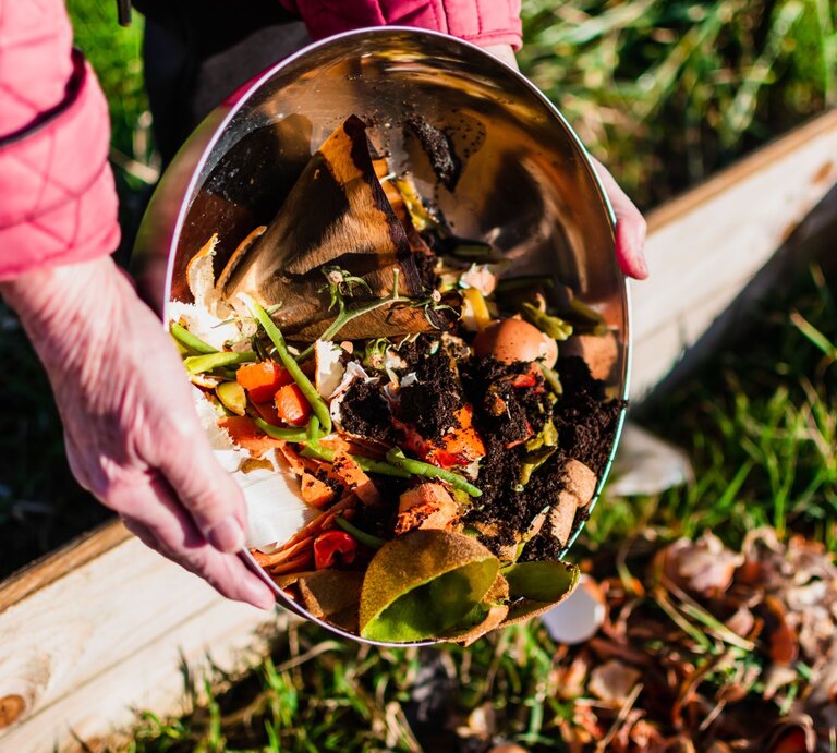 Eine Person in rosa Jacke entleert eine Schüssel mit Gemüseabfällen und Erde auf ein Beet im sonnigen Garten. | © Shutterstock/Reflexpixel