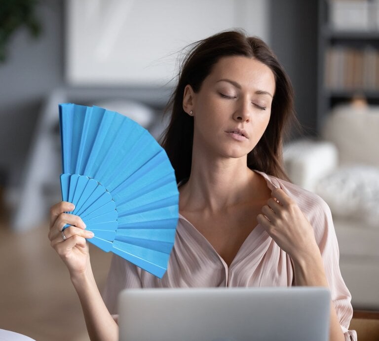 Das Bild zeigt eine Frau, die an einem Laptop sitzt und sich mit einem blauen Fächer Luft zufächelt. Sie wirkt erschöpft. | © iStock.com/fizkes