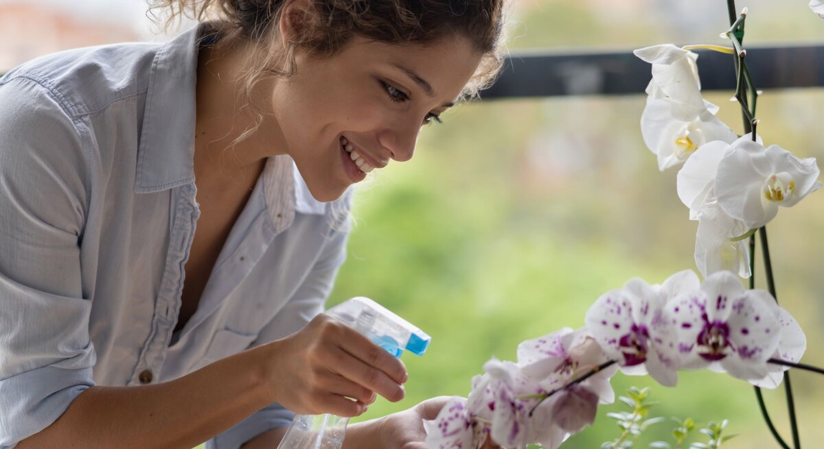 Eine junge Frau mit lockigem Haar sprüht lächelnd Wasser auf weiße Orchideen vor einem unscharfen grünen Hintergrund.