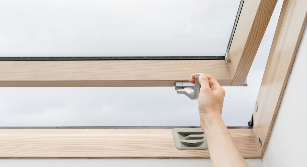 Eine Hand öffnet ein Dachfenster aus Holz mit einem grauen Griff an einem bewölkten Tag.
