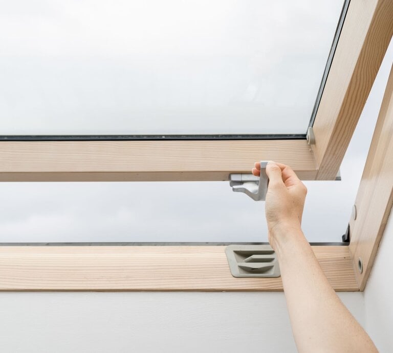 Eine Hand öffnet ein Dachfenster aus Holz mit einem grauen Griff an einem bewölkten Tag. | © AdobeStock/brizmaker