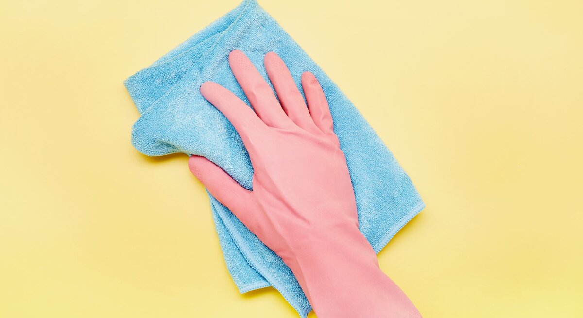 Eine Hand im rosa Gummihandschuh wischt mit einem blauen Microfaser Putzlappen über einen gelben Hintergrund.