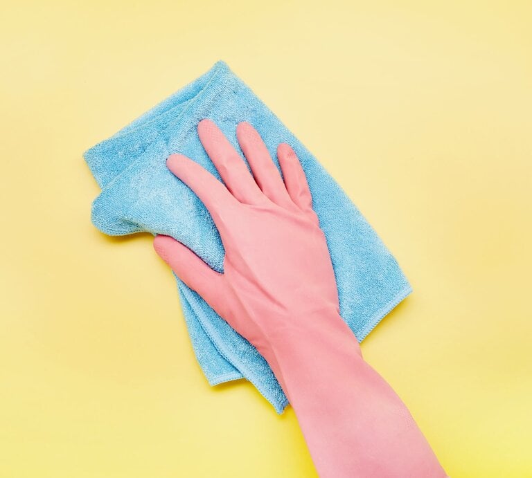 Eine Hand im rosa Gummihandschuh wischt mit einem blauen Microfaser Putzlappen über einen gelben Hintergrund. | © gettyimages/	the_burtons