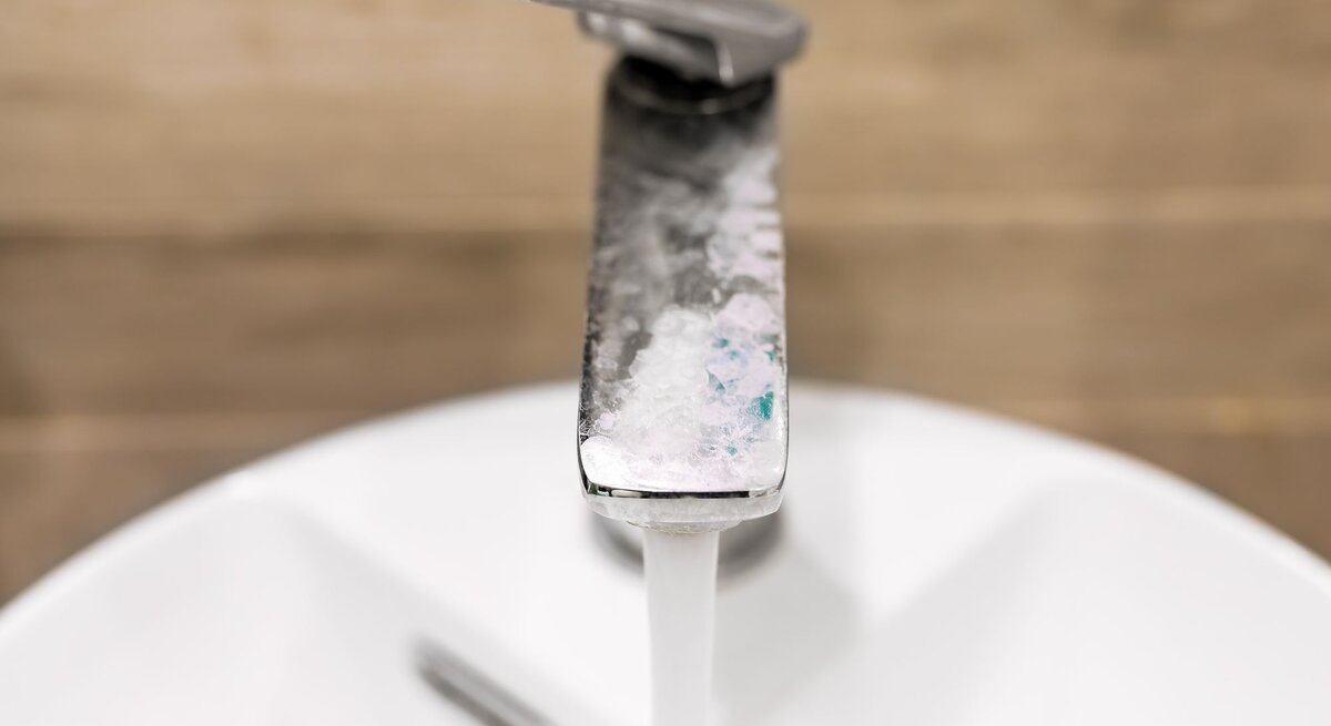 Nahaufnahme eines Wasserhahns mit Kalkablagerungen, aus dem Wasser in ein weißes Waschbecken fließt, vor unscharfem Holzhintergrund.