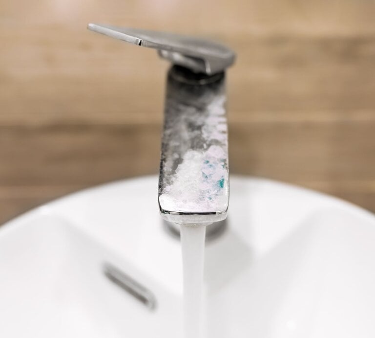 Nahaufnahme eines Wasserhahns mit Kalkablagerungen, aus dem Wasser in ein weißes Waschbecken fließt, vor unscharfem Holzhintergrund. | © GettyImages/valiantsin suprunovich