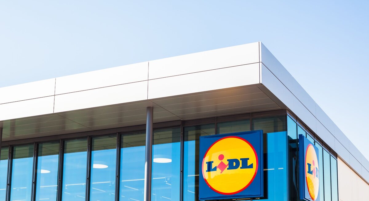  Das ist ein Bild eines modernen Gebäudes mit einer großen Glasfassade. Auf der Fassade sind zwei große, quadratische Schilder mit dem Logo von Lidl, einer bekannten Supermarktkette, zu sehen. Das Logo besteht aus einem blauen Hintergrund mit einem gelben Kreis, in dem sich der Name "Lidl" in roten Buchstaben befindet. Darüber ist ein kleiner, stilisierter Kopf mit einem gelben Umriss abgebildet. Das Gebäude selbst hat eine klare, rechtwinklige Form mit einem flachen Dach, was typisch für kommerzielle Gebäude dieser Art ist.