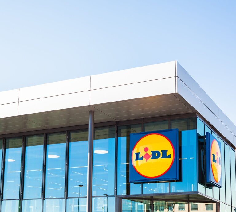  Das ist ein Bild eines modernen Gebäudes mit einer großen Glasfassade. Auf der Fassade sind zwei große, quadratische Schilder mit dem Logo von Lidl, einer bekannten Supermarktkette, zu sehen. Das Logo besteht aus einem blauen Hintergrund mit einem gelben Kreis, in dem sich der Name "Lidl" in roten Buchstaben befindet. Darüber ist ein kleiner, stilisierter Kopf mit einem gelben Umriss abgebildet. Das Gebäude selbst hat eine klare, rechtwinklige Form mit einem flachen Dach, was typisch für kommerzielle Gebäude dieser Art ist. | © AdobeStock/ dvoevnore