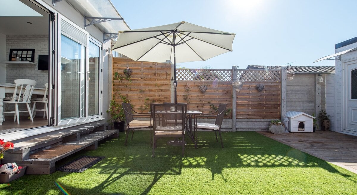 Sonnige Terrasse mit grüner Kunstrasenfläche, Tisch und Stühlen unter einem großen Sonnenschirm, daneben ein offenes Wohnzimmer.