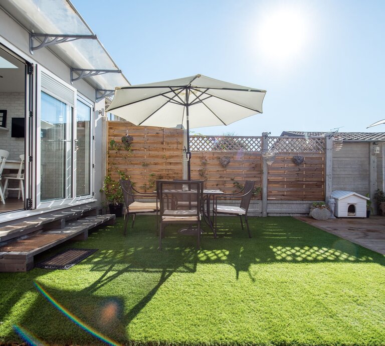 Sonnige Terrasse mit grüner Kunstrasenfläche, Tisch und Stühlen unter einem großen Sonnenschirm, daneben ein offenes Wohnzimmer. | © Getty Images/John Keeble