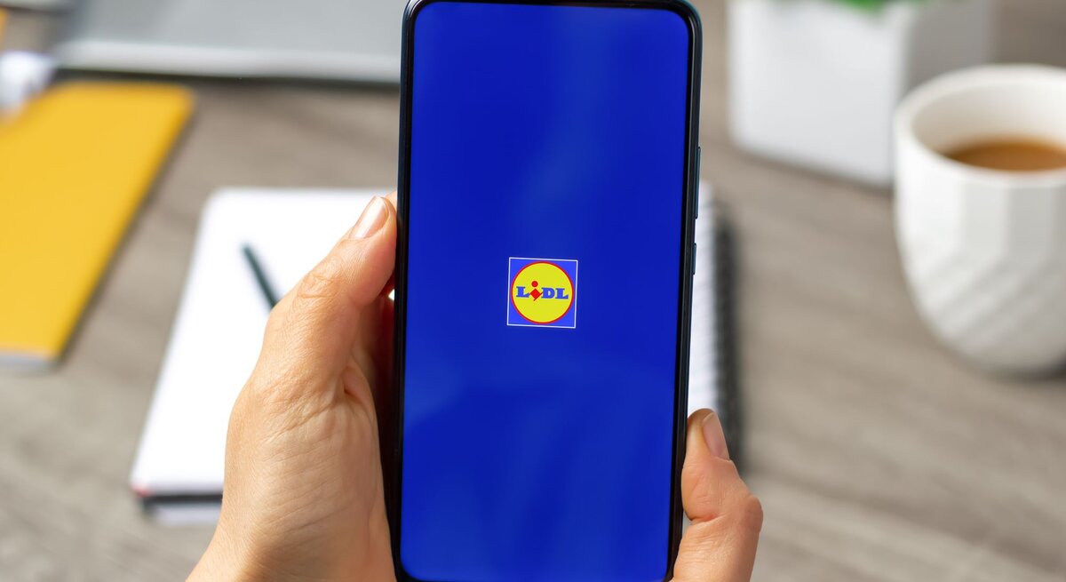 Eine Hand hält ein Smartphone mit Lidl-Logo auf blauem Bildschirm, im Hintergrund unscharfe Büroelemente.