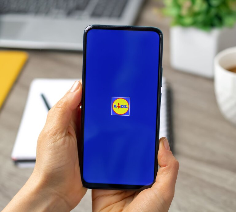 Eine Hand hält ein Smartphone mit Lidl-Logo auf blauem Bildschirm, im Hintergrund unscharfe Büroelemente. | © AdobeStock/ Oleksandr