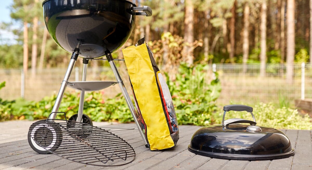 Schwarzer Kugelgrill bereit zum Anfeuern auf Terrasse mit Tüte Holzkohle, im Hintergrund Kiefernwald.