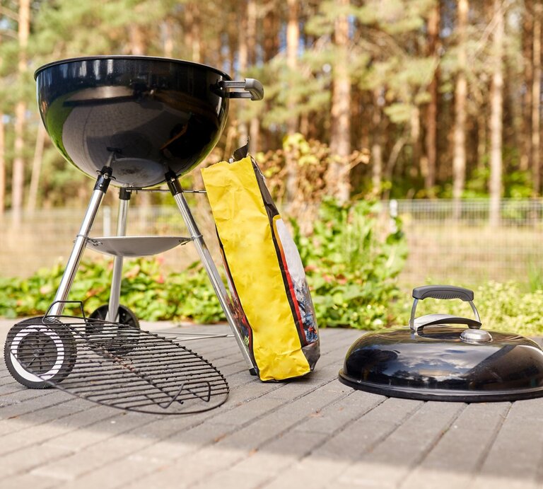 Schwarzer Kugelgrill bereit zum Anfeuern auf Terrasse mit Tüte Holzkohle, im Hintergrund Kiefernwald. | © Adobe Stock/Syda Productions