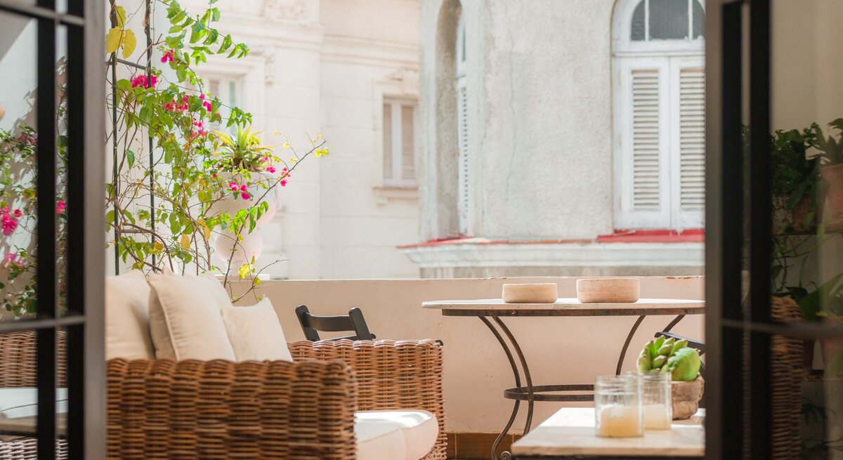 Eine sonnige Altbau-Loggia mit Korbsessel, weißen Polstern und kleinem Tisch, umrankt von pink blühender Bougainvillea.