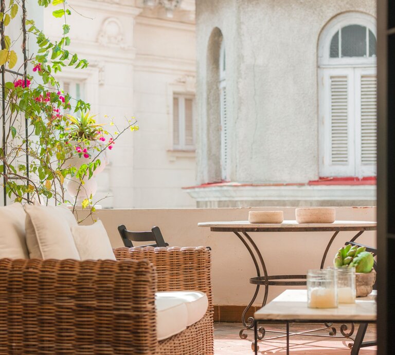 Eine sonnige Altbau-Loggia mit Korbsessel, weißen Polstern und kleinem Tisch, umrankt von pink blühender Bougainvillea. | © Shutterstock/Wirestock Creators