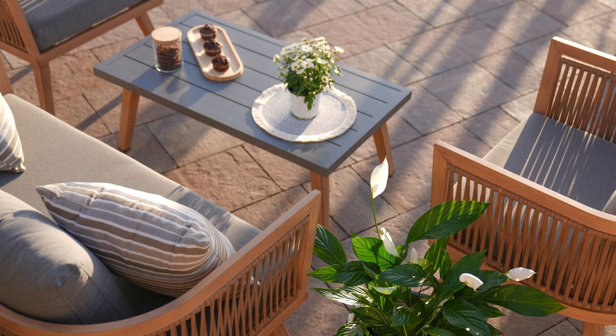 Elegante Gartenlounge mit Holzmöbeln und grauen Polstern um einen Tisch mit Muffins, Blumen und Keksdose in der Abendsonne.