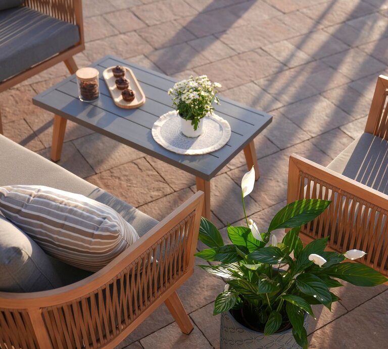 Elegante Gartenlounge mit Holzmöbeln und grauen Polstern um einen Tisch mit Muffins, Blumen und Keksdose in der Abendsonne. | © Adobe Stock/ Ahmad TB