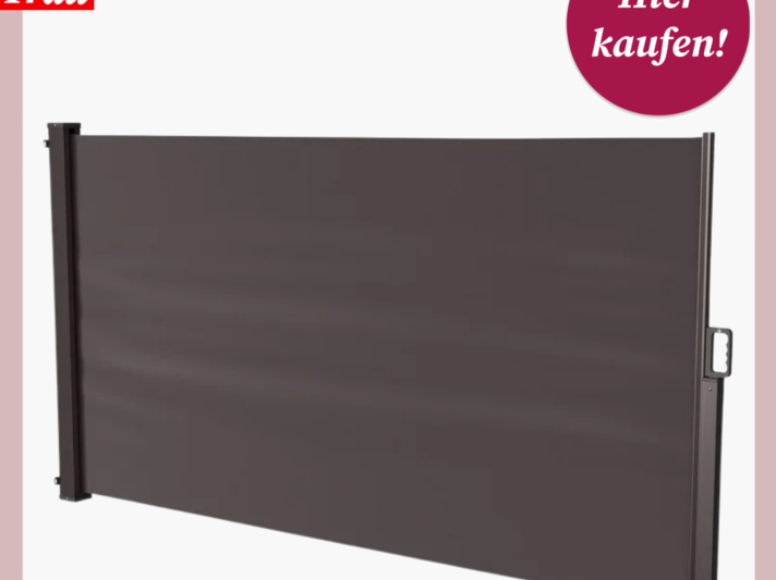  Das Bild zeigt einen ausziehbaren Sichtschutz, der aufrecht montiert ist. Der Sichtschutz scheint aus einem robusten, einfarbigen Material, wahrscheinlich Stoff oder Kunststoff, zu bestehen und ist vollständig ausgezogen. Die Konstruktion ist in einem schlichten, dunklen Farbton gehalten. Links ist eine Kassette zu sehen, in der der Sichtschutz aufgerollt wird, und rechts ist das Ende des Sichtschutzes mit einem Griff versehen, der vermutlich zum Ausziehen und Einziehen dient. Oben auf dem Bild befindet sich ein Logo mit dem Text "Bild der Frau" und ein kreisförmiges Etikett mit der Aufschrift "Hier kaufen!", was darauf hindeutet, dass das Bild für Werbe- oder Verkaufszwecke verwendet wird. | © Lidl 