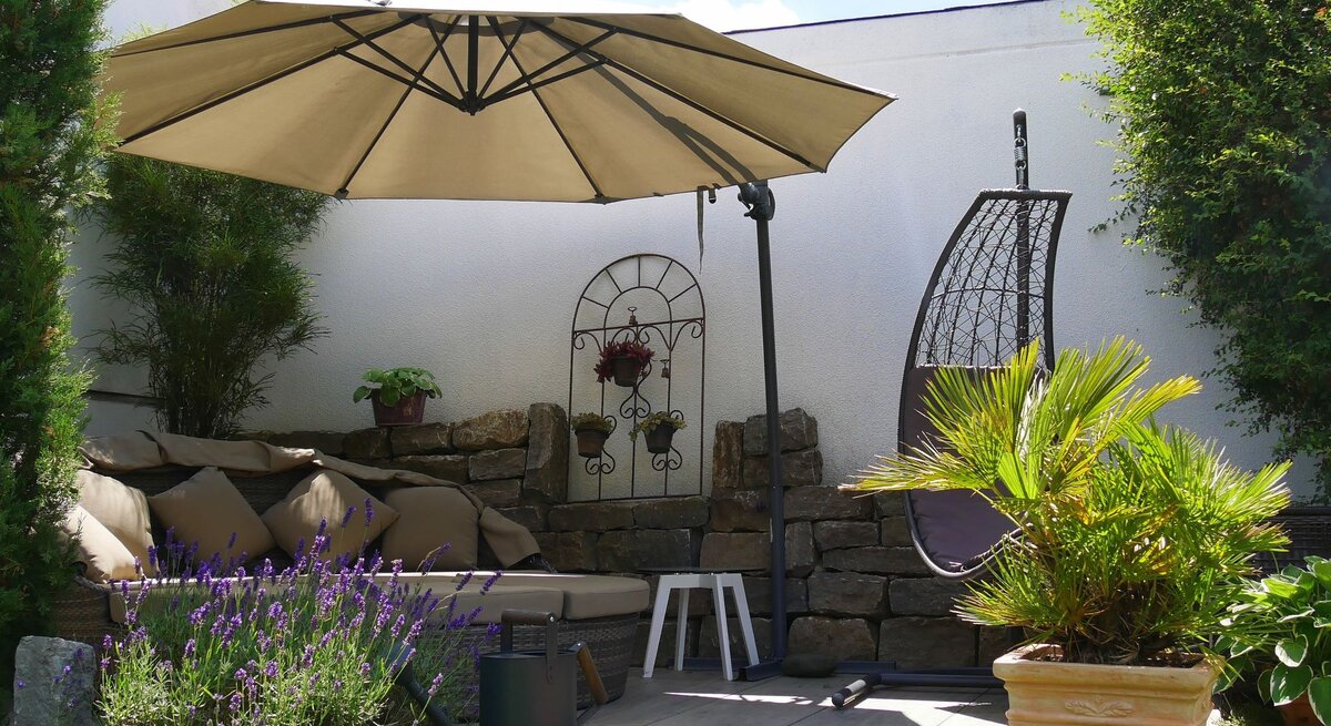 Gemütliche Gartenecke mit Sonnenliege, Hängesessel, Sonnenschirm und blühendem Lavendel vor heller Mauer und Steinwand.