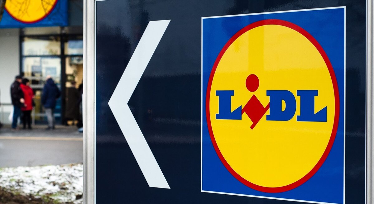  Das Bild zeigt ein großes Schild mit einem Pfeil und dem Logo des Supermarkts Lidl. Der Pfeil weist nach links, was darauf hinweist, dass die Filiale in diese Richtung liegt. Im Hintergrund sieht man den Eingangsbereich eines Lidl-Marktes mit einigen Personen, die sich in der Nähe befinden. Der Boden ist teilweise mit Schnee bedeckt, was auf winterliche Wetterbedingungen schließen lässt.