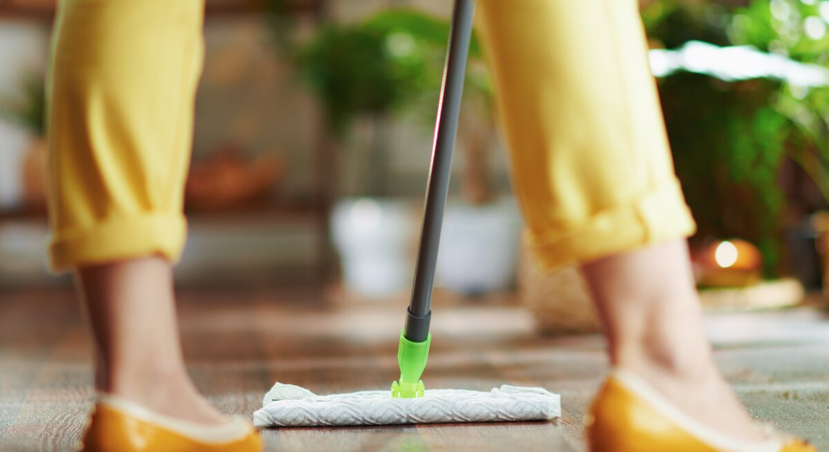  Das Bild zeigt die untere Hälfte einer Person beim Bodenwischen. Die Person trägt gelbe Hosen und passende gelbe Schuhe, was auf ein farblich abgestimmtes Outfit hinweist. Sie benutzt einen Swiffer Bodenwischer mit einem feuchten Bodentuch, das am Ende des Wischers angebracht ist, um den Holzboden zu reinigen. Die Szene spielt sich in einem hellen Raum mit Pflanzen im Hintergrund ab, was eine lebendige und gemütliche häusliche Umgebung andeutet. Der Fokus liegt auf der Wischaktion und dem Wischmop, was die einfache und effiziente Reinigung von Böden mit diesen modernen Reinigungswerkzeugen unterstreicht.