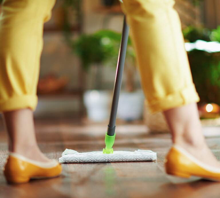  Das Bild zeigt die untere Hälfte einer Person beim Bodenwischen. Die Person trägt gelbe Hosen und passende gelbe Schuhe, was auf ein farblich abgestimmtes Outfit hinweist. Sie benutzt einen Swiffer Bodenwischer mit einem feuchten Bodentuch, das am Ende des Wischers angebracht ist, um den Holzboden zu reinigen. Die Szene spielt sich in einem hellen Raum mit Pflanzen im Hintergrund ab, was eine lebendige und gemütliche häusliche Umgebung andeutet. Der Fokus liegt auf der Wischaktion und dem Wischmop, was die einfache und effiziente Reinigung von Böden mit diesen modernen Reinigungswerkzeugen unterstreicht. | © Adobe Stock/Alliance