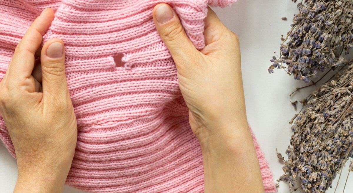 Hände halten rosa gestrickten Pullover neben getrocknetem Lavendel auf weißem Untergrund.