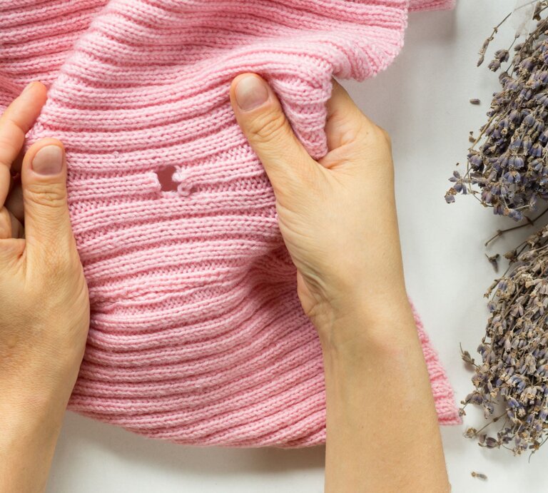 Hände halten rosa gestrickten Pullover neben getrocknetem Lavendel auf weißem Untergrund. | © Gettyimages/Tatiana Foxy