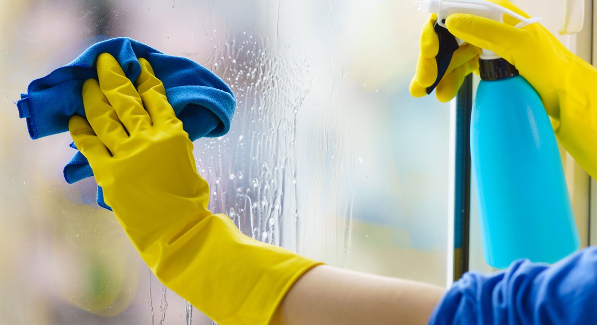 Behandschuhte Hände putzen Fenster mit Sprühflasche und Lappen