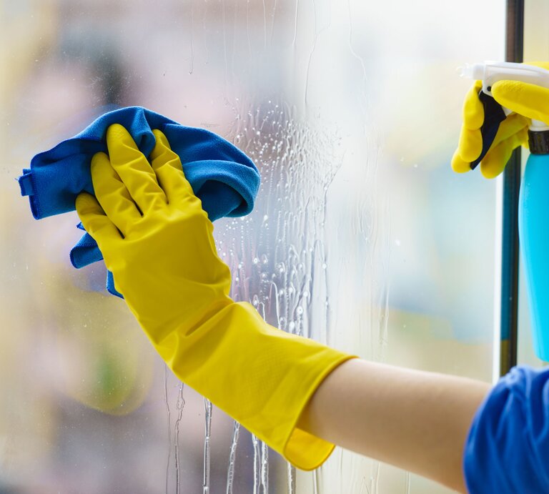 Behandschuhte Hände putzen Fenster mit Sprühflasche und Lappen | © Adobe Stock