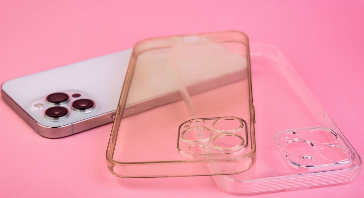 Eine silberne Smartphone-Rückseite mit drei Kameralinsen liegt auf rosa Hintergrund, daneben zwei transparente Handyhüllen, eine davon ist vergilbt.