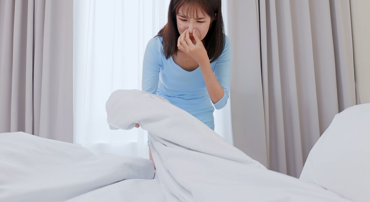 Eine Frau hält sich die Nase zu, während Sie Bettwäsche auf dem Bett hochhebt.
