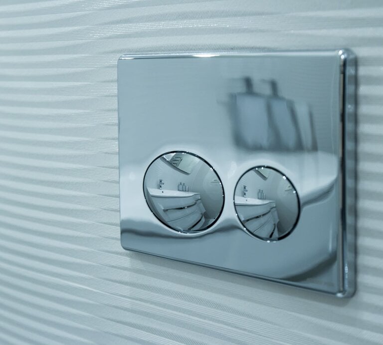 Verchromtes WC-Spülsystem mit zwei runden Tasten spiegelt ein modernes Badezimmer, montiert auf geriffelter weißer Wand. | © Adobe Stock/Igor Gromoff