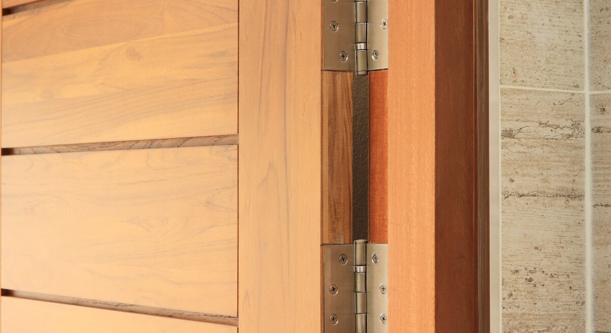 Holztür mit sichtbarem Metallscharnier, das an einer beige gekachelten Wand montiert ist.