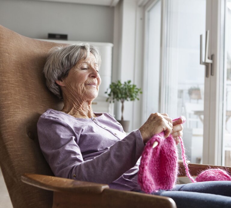 Eine ältere Frau sitzt lächelnd in einem Sessel und strickt. Sie genießt die Wärme in der Wohnung. | © getty images/ Westend61
