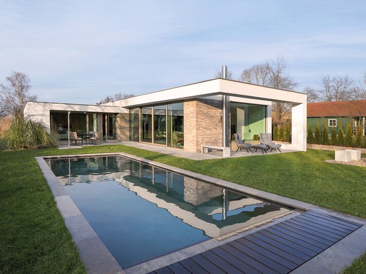 Modernes Flachdachhaus mit großen Glasfronten und Natursteinfassade, davor ein schmaler Pool und gepflegter Rasen bei Sonnenschein. | © LOTTO24