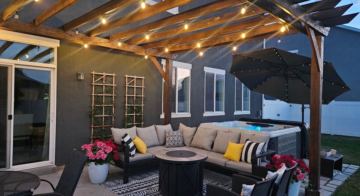 Gemütliche überdachte Terrasse am Abend mit Lichterketten, Lounge-Sofa, dekorativen Kissen, Feuertisch und beleuchtetem Whirlpool im Hintergrund.