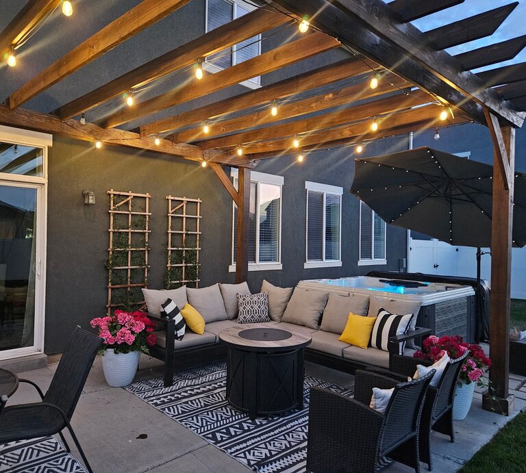 Gemütliche überdachte Terrasse am Abend mit Lichterketten, Lounge-Sofa, dekorativen Kissen, Feuertisch und beleuchtetem Whirlpool im Hintergrund. | © Shutterstock/ LindsayNorton