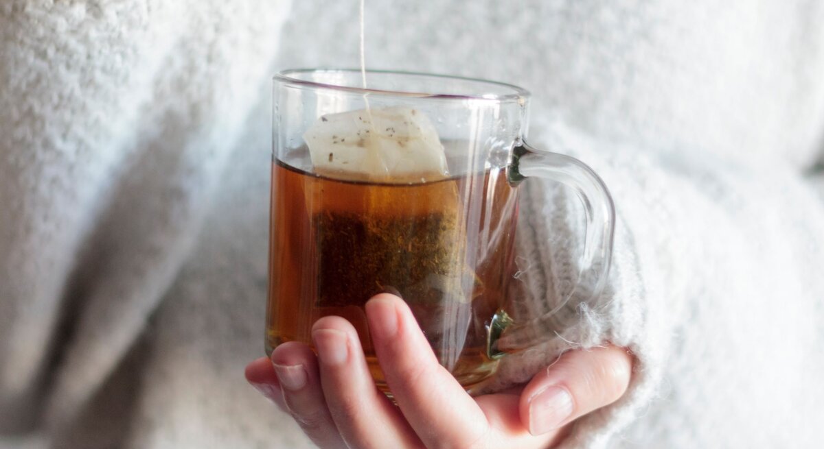 Eine Hand in einem flauschigen weißen Pullover hält eine Tasse heißen Tee, während ein Teebeutel langsam zieht.