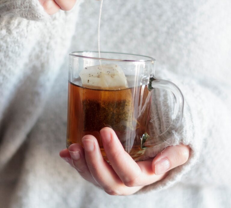 Eine Hand in einem flauschigen weißen Pullover hält eine Tasse heißen Tee, während ein Teebeutel langsam zieht. | © AdobeStock/ raquel