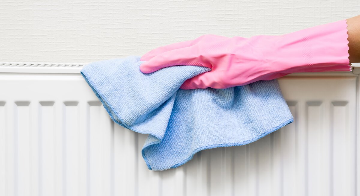 Eine Hand in pinkem Gummihandschuh wischt mit einem blauen Mikrofasertuch einen weißen Heizkörper vor cremefarbener Wand ab.