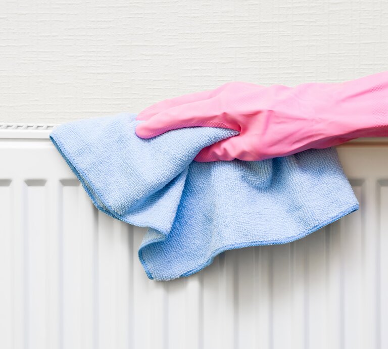 Eine Hand in pinkem Gummihandschuh wischt mit einem blauen Mikrofasertuch einen weißen Heizkörper vor cremefarbener Wand ab. | © Adobe Stock/ fotoduets