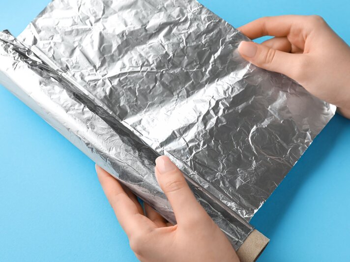 Zwei Hände, die eine Rolle Aluminiumfolie abrollen. Die Aluminiumfolie ist silbern und leicht zerknittert, und sie wird gegen einen blauen Hintergrund gehalten. | © Adobe Stock / Pixel-Shot